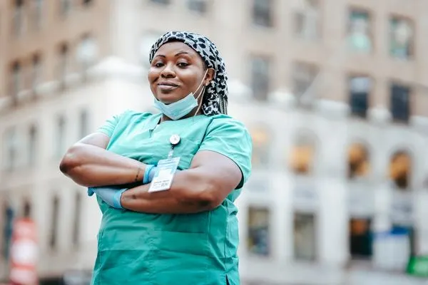 lady nurse in greed scrubs and name badge crossing her arms outdoors in front of building