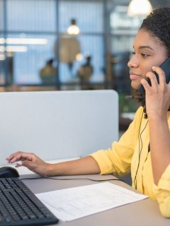 featured woman sitting in front of computer at work desk talking on the phone to as a nurse