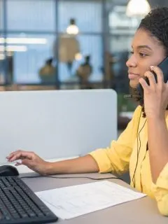featured woman sitting in front of computer at work desk talking on the phone to as a nurse