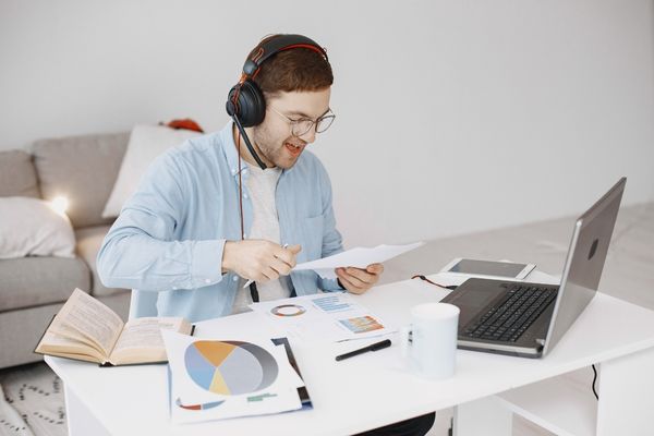 man working from home with headset, sitting in front of laptop while hold a paper that he's reading