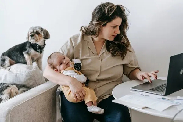 nursing student working from home on kitchen table laptop while holding baby and puppy in the background