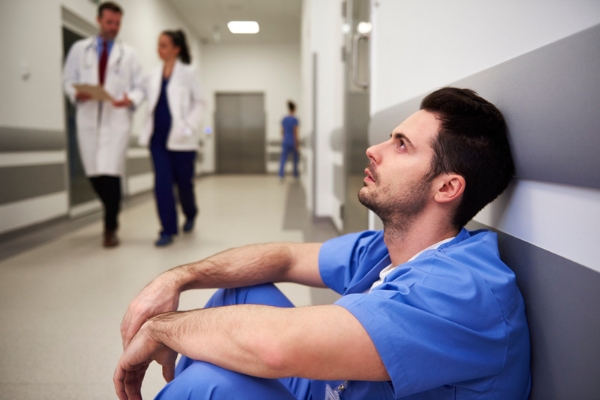 a depressed male nurse sitting against the wall while medical staff walks around him at the hospital